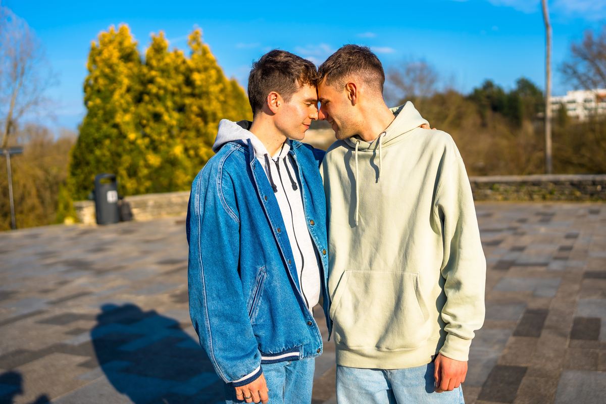 Two young men, one in a denim jacket and one in a pale hoodie, touching foreheads in a sunny park