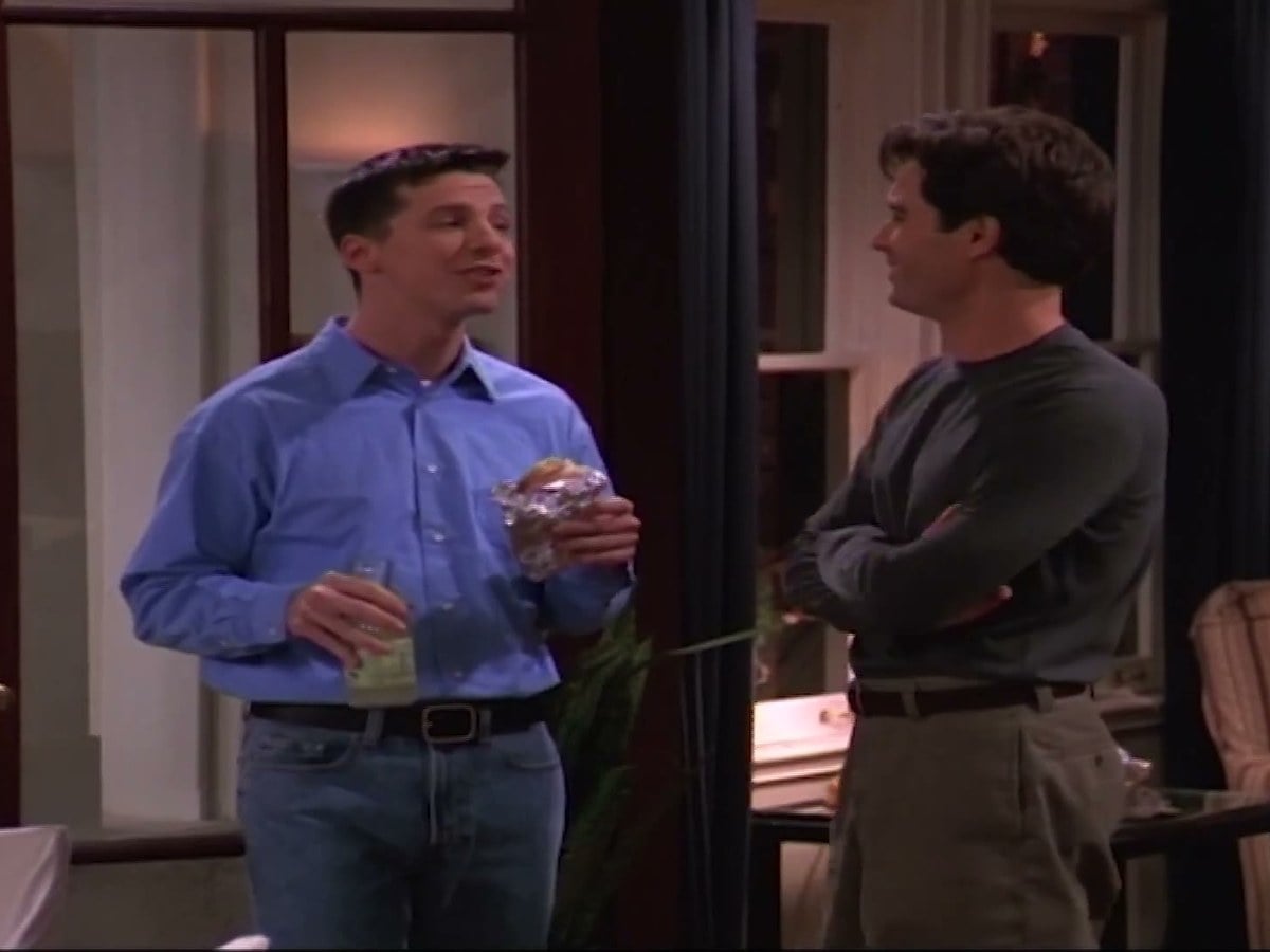 Sean Hayes as Jack, in a blue shirt holding a drink, talking to a smiling Eric McCormack as Will, arms crossed
