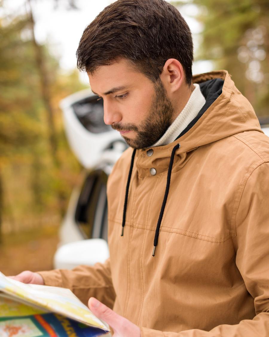 A bearded man in a tan jacket reading a paper map beside his car