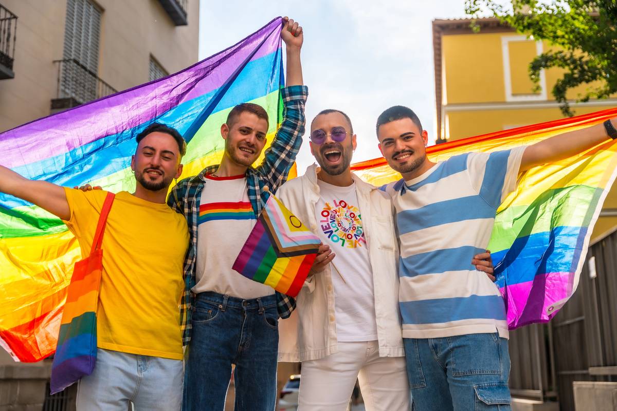 Four smiling men with their arms around each other under large rainbow flags on a city street at Pride