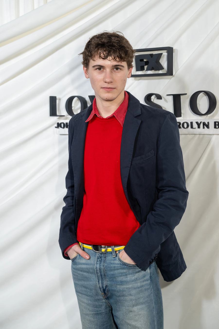 Sam Nivola in a red sweater, navy blazer and jeans at the FX Love Story premiere in New York