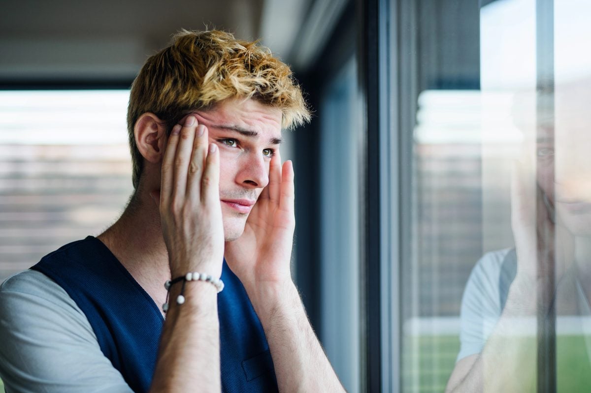 A young man with blond hair holding his head in his hands beside a window