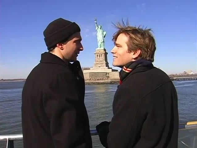 Van Hansis and Jake Silbermann in winter coats facing each other on a ferry with the Statue of Liberty behind them in As the World Turns