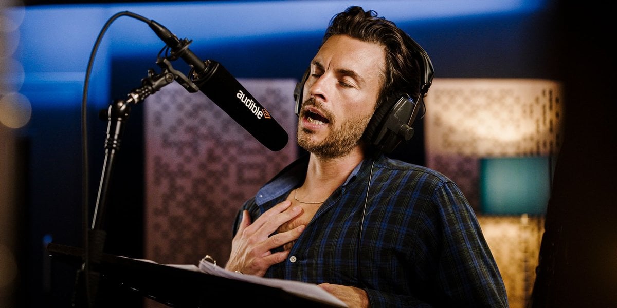 Jonathan Bailey with eyes closed and a hand on his chest, recording at an Audible microphone