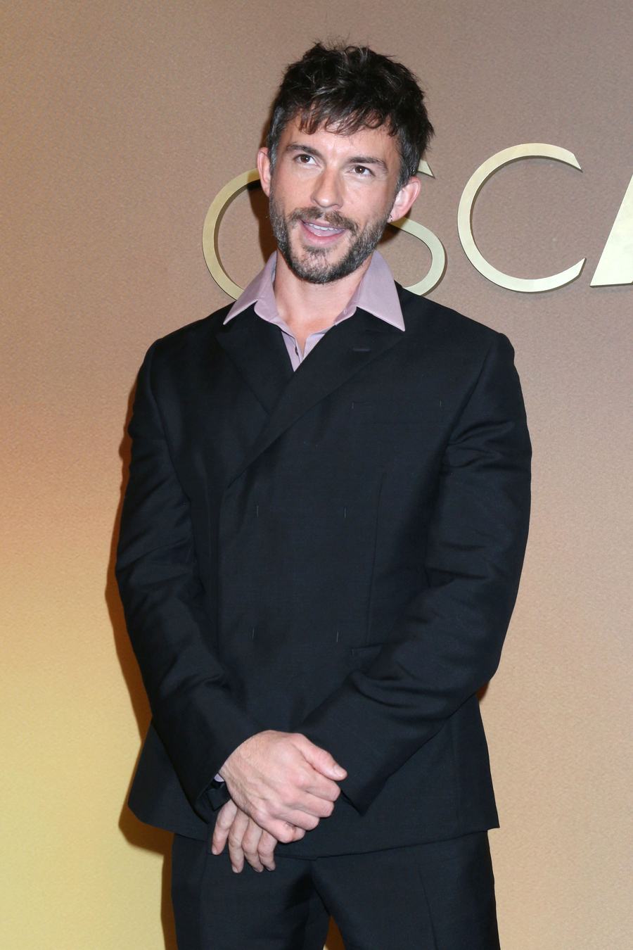 Jonathan Bailey smiling in a black suit and pale pink shirt