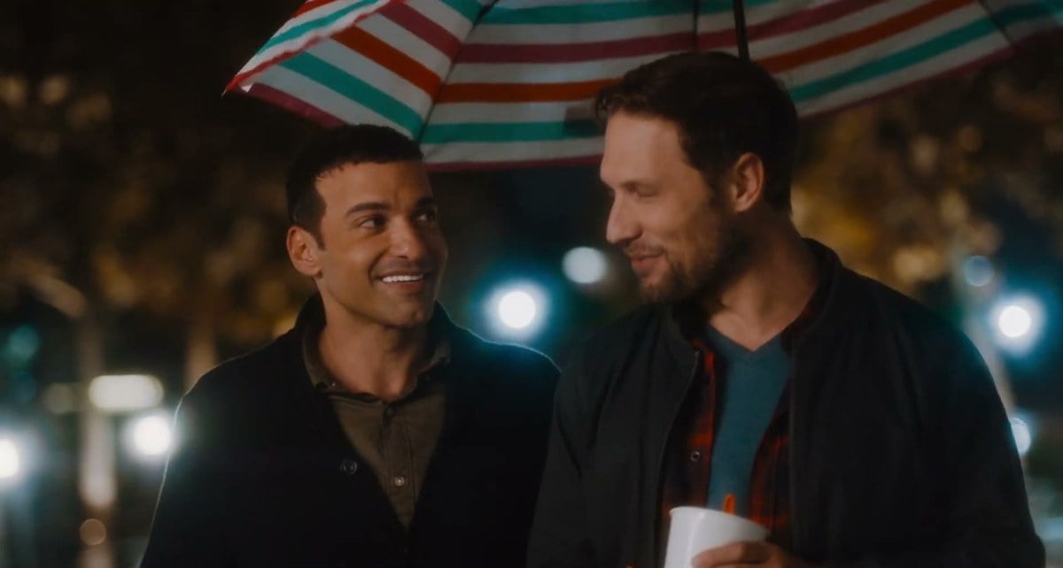 Haaz Sleiman and Michael Cassidy smiling at each other under a striped umbrella at night in Breaking Fast