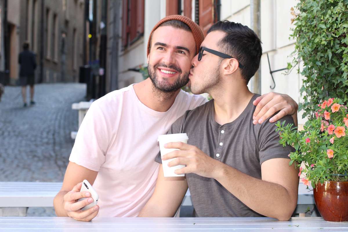 A man in glasses kisses his smiling boyfriend in a beanie on the cheek at an outdoor cafe table