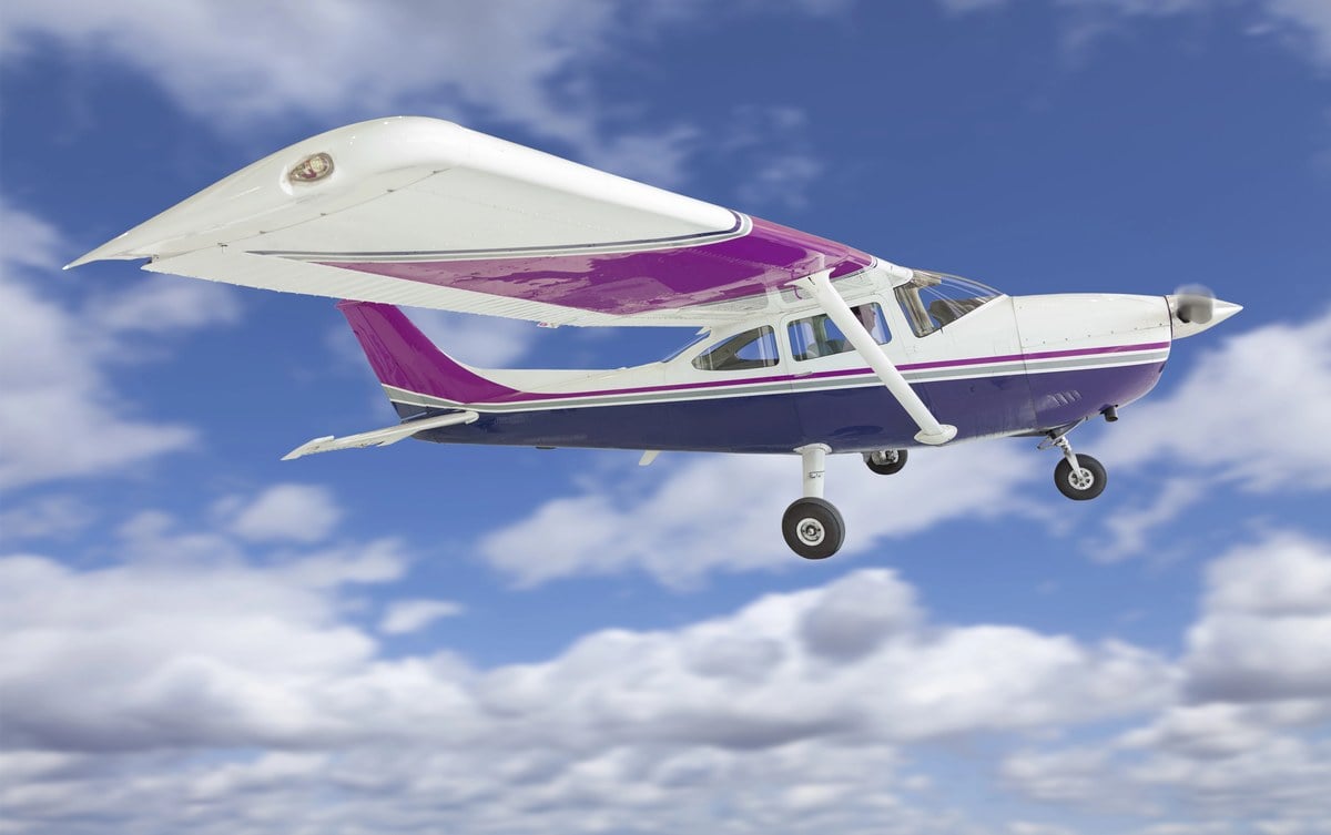 A white and purple Cessna 172 single-propeller plane flying through a blue sky with clouds