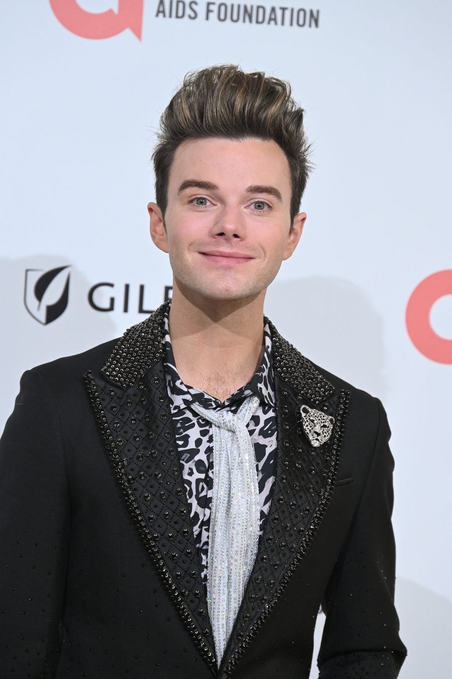Chris Colfer smiling in a black embellished blazer and leopard-print shirt