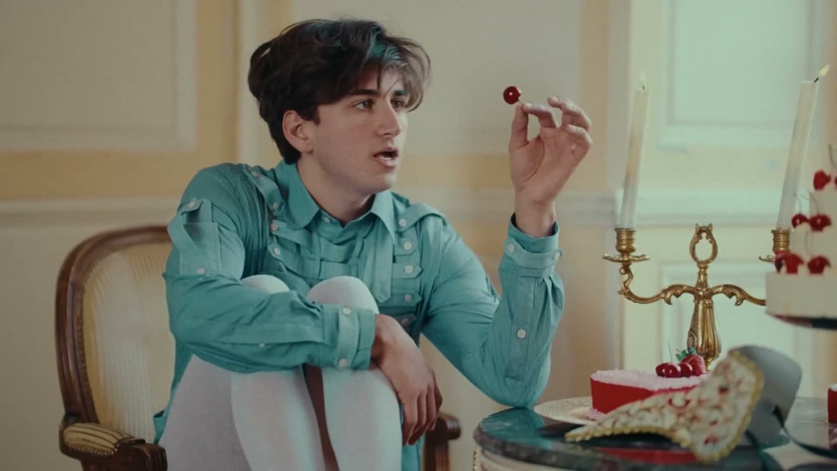 Sebastian Croft in a teal shirt holding up a cherry in an ornate room