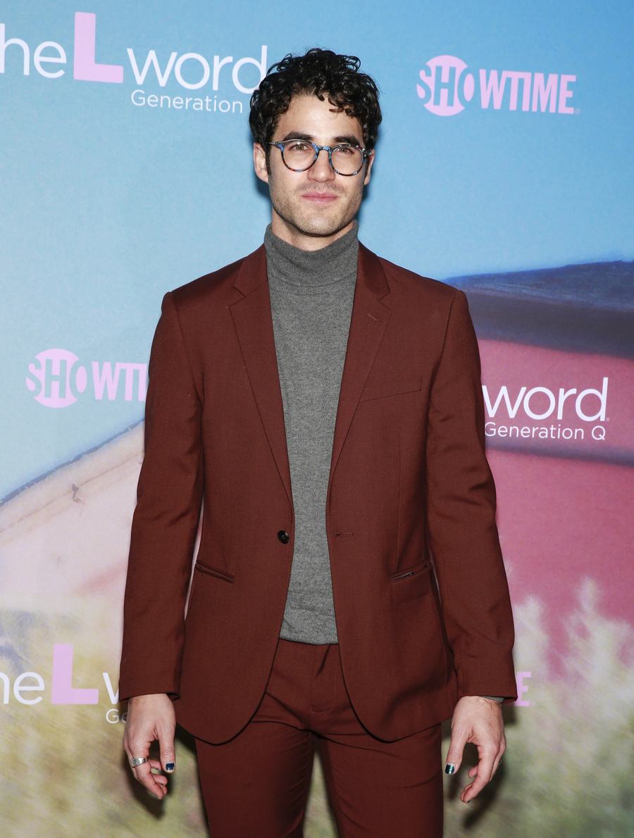 Darren Criss in glasses, a rust-colored suit and grey turtleneck on a red carpet