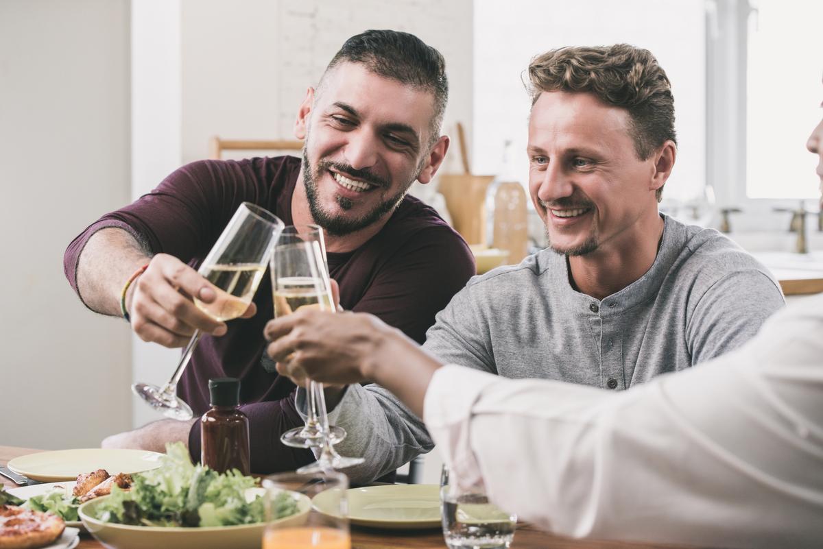Two laughing men clinking champagne glasses at a dinner table with friends