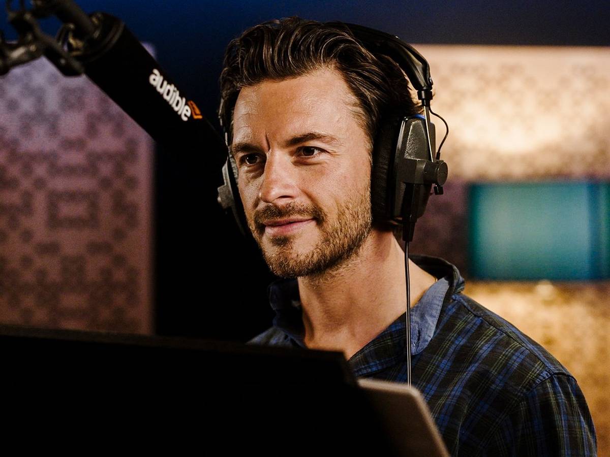Jonathan Bailey in headphones and an open plaid shirt, smiling at the microphone in a recording booth