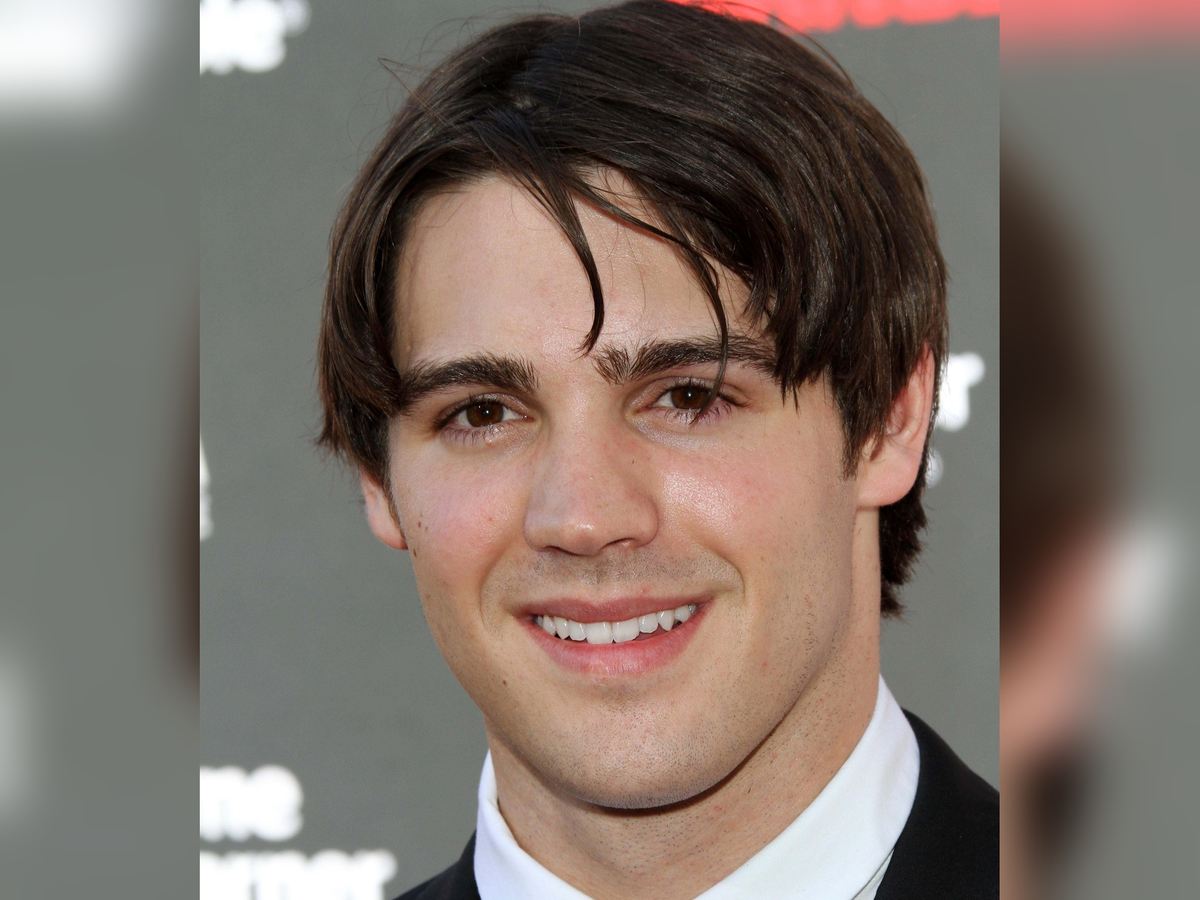 Steven R. McQueen smiling in a black suit and white shirt on a red carpet