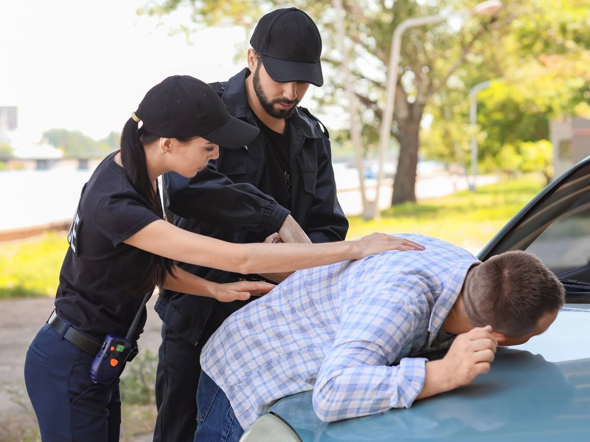 Two police officers arresting a man who is bent over the hood of a car