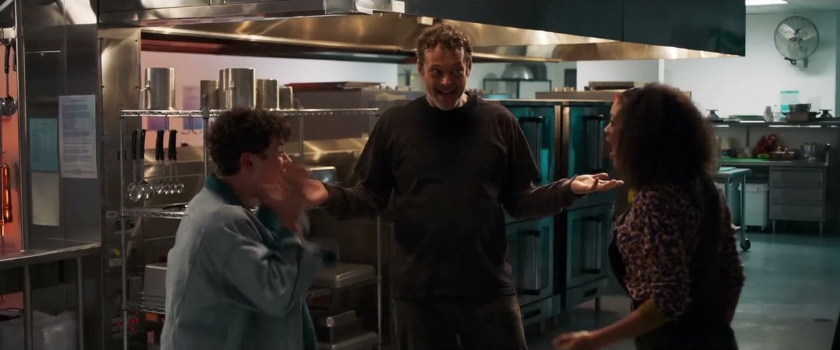 Vince Vaughn grinning with his arms spread in a school kitchen as Misha Osherovich and Celeste O'Connor react