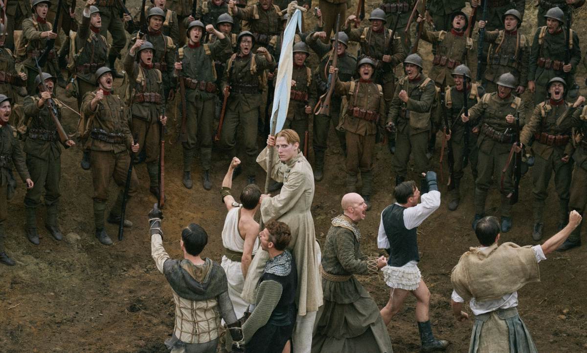Soldiers in costume put on a show in a field, watched by a crowd of troops, in Coward