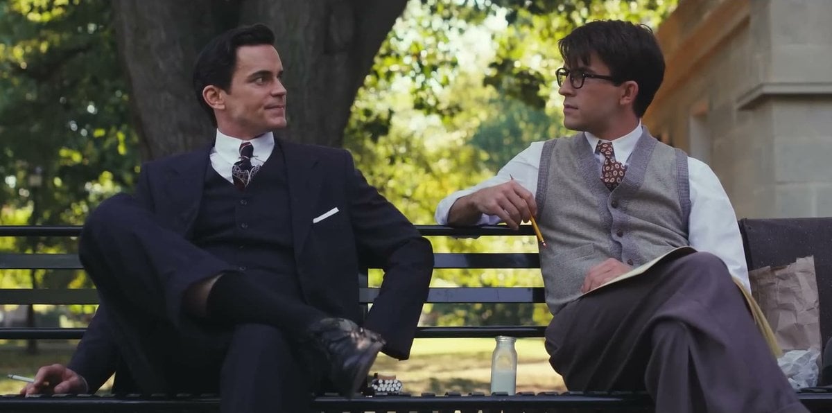 Matt Bomer in a navy three-piece suit and Jonathan Bailey in glasses and a sweater vest smiling at each other on a park bench in Fellow Travelers