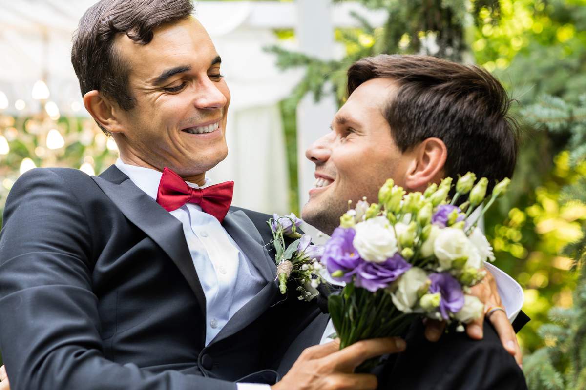 Two smiling grooms embracing at a garden wedding, one holding a bouquet