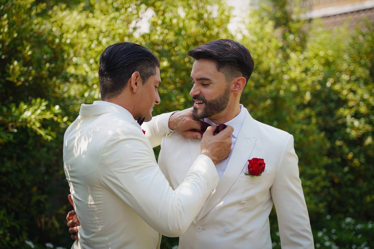 Two grooms in white suits at a garden wedding, one straightening the other's bow tie