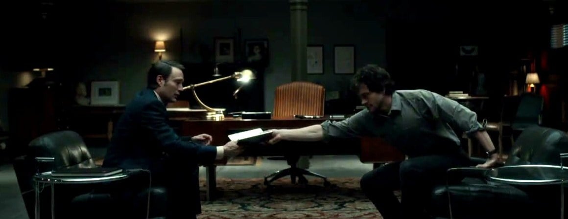 Mads Mikkelsen as Hannibal Lecter taking a notebook from Hugh Dancy's Will Graham across his office during a therapy session