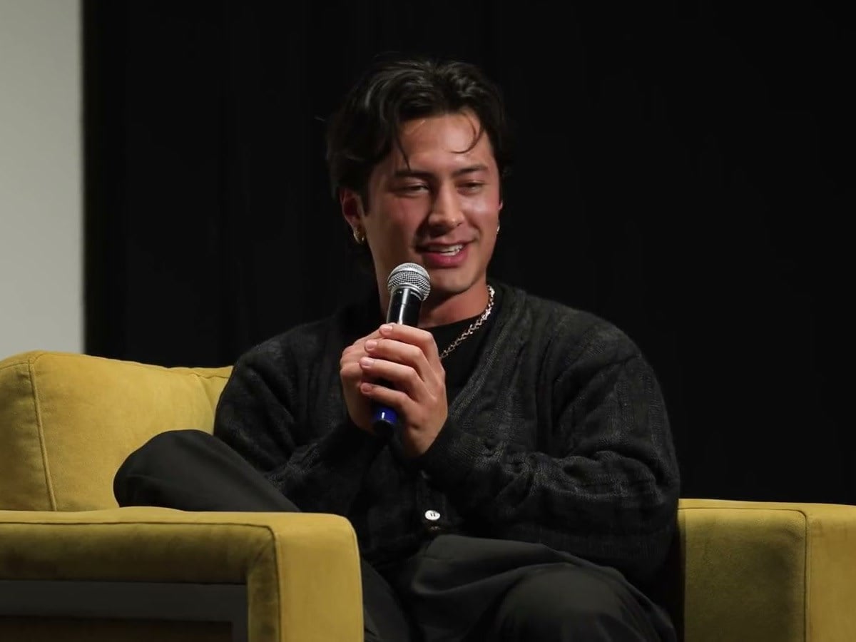Hudson Williams smiling and holding a microphone while seated on stage in a dark sweater