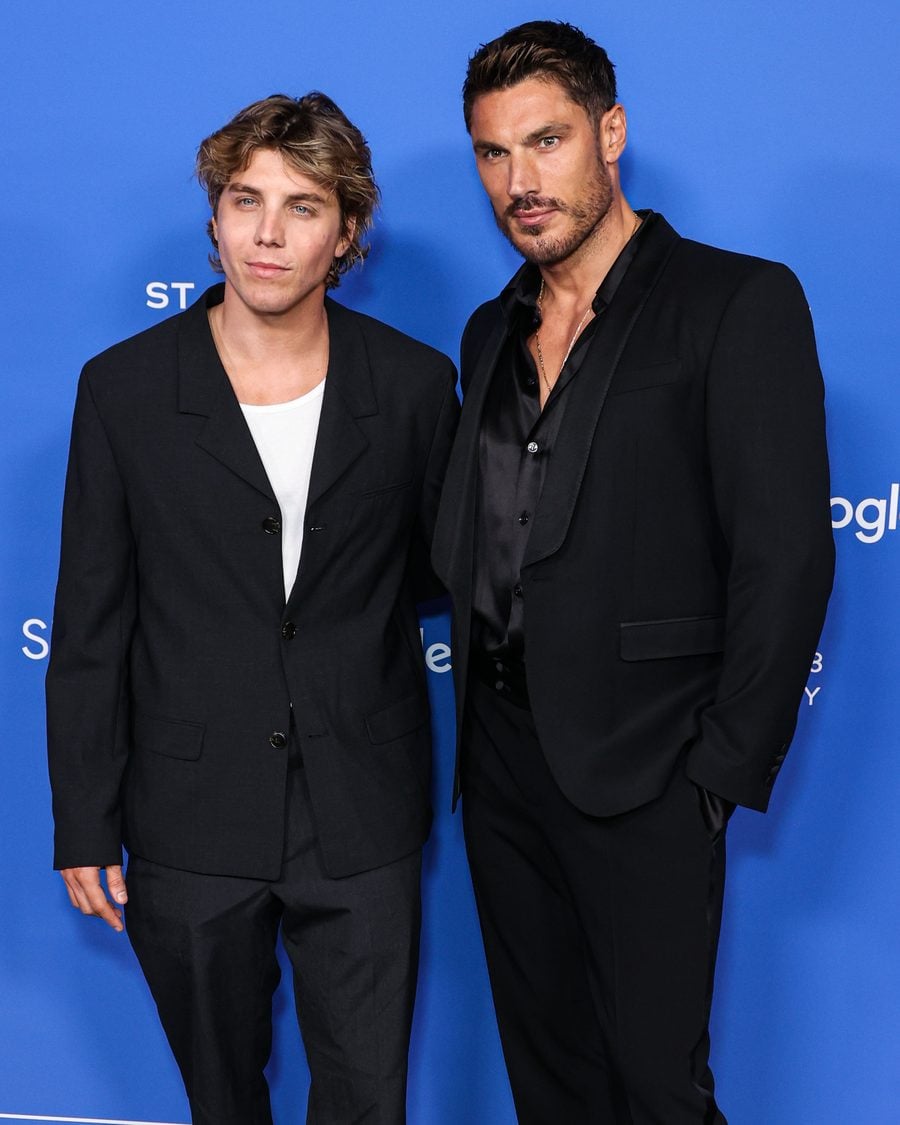 Lukas Gage and Chris Appleton in black suits on a blue red-carpet backdrop
