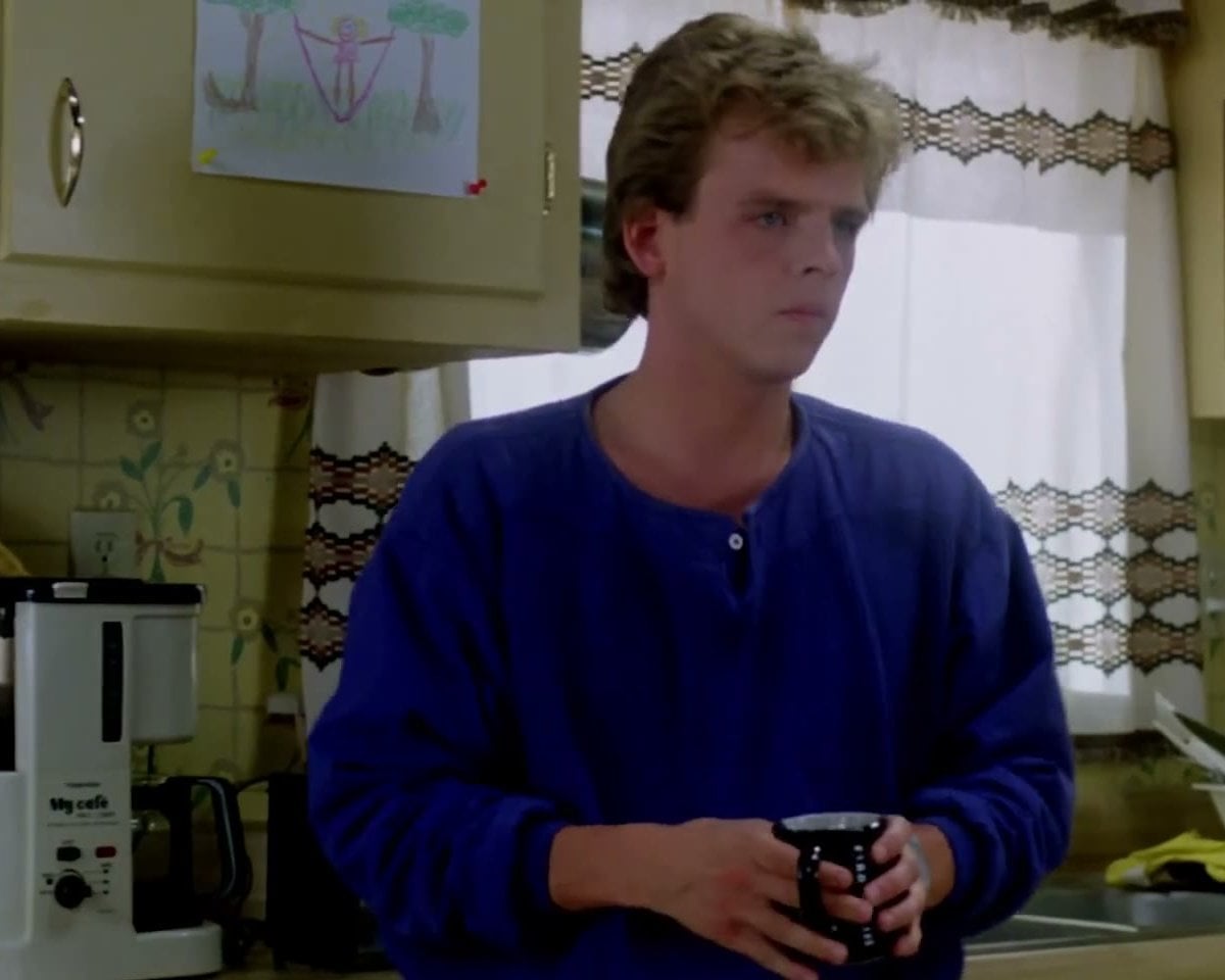 Mark Patton as Jesse, in a blue sweater holding a coffee mug in the family kitchen