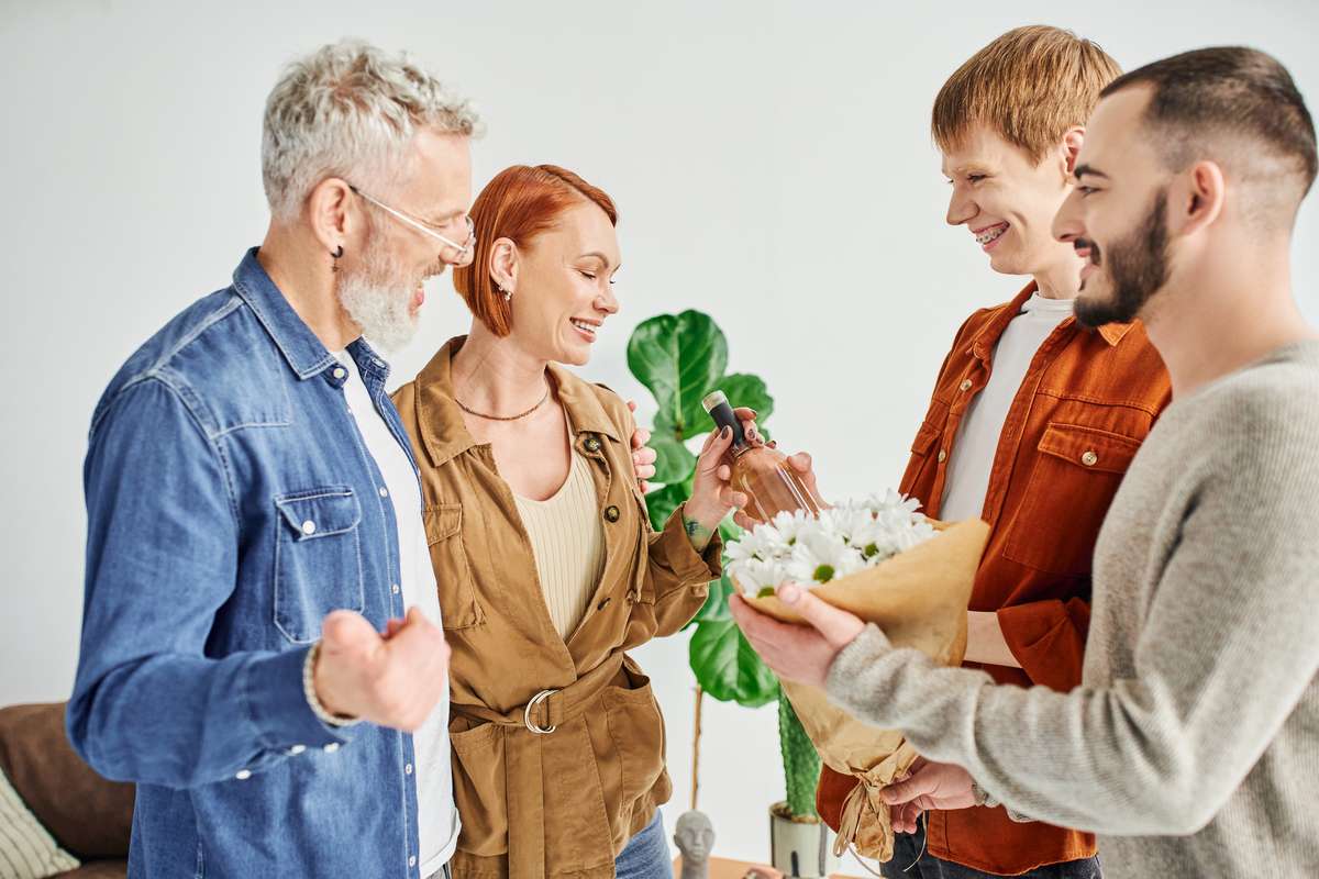A smiling young couple of two men handing flowers and wine to an older man and woman at their home