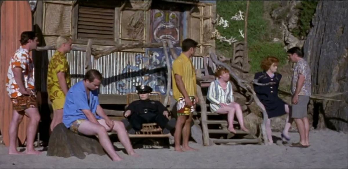 The surfers and beach girls of Psycho Beach Party gathered outside a beach shack, with a police officer seated in the middle