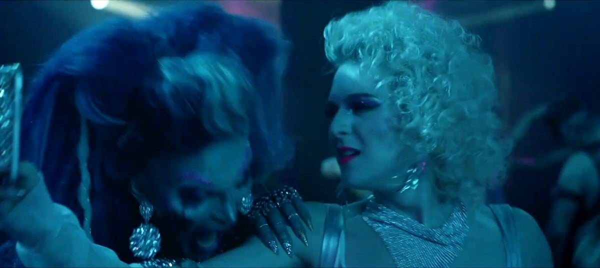 Two drag queens in big blue and platinum wigs and sparkling costumes on a club dance floor in Queens of the Dead