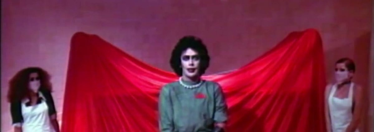 Tim Curry as Dr. Frank-N-Furter in a green lab coat and pearls, in front of a red sheet, with Magenta and Columbia beside him