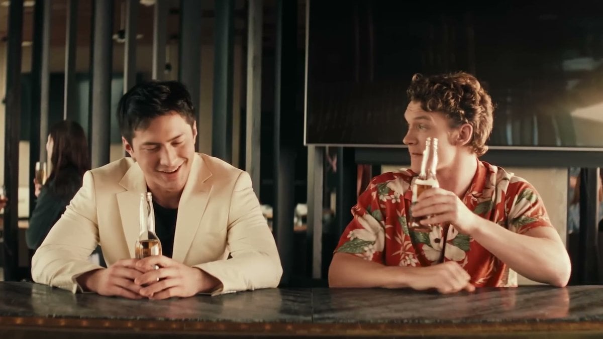 Hudson Williams and Connor Storrie as Shane and Ilya, sitting at a bar with beers and smiling