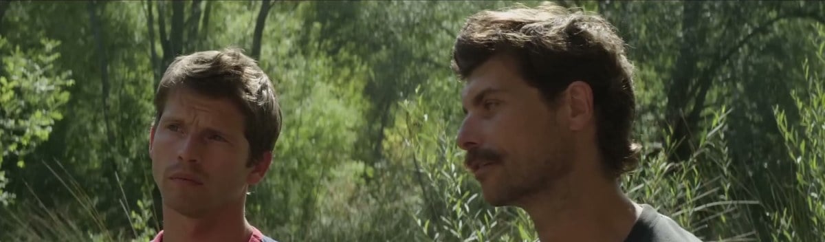 Pierre Deladonchamps and Christophe Paou, both in T-shirts, standing among the trees by the lake in Stranger by the Lake
