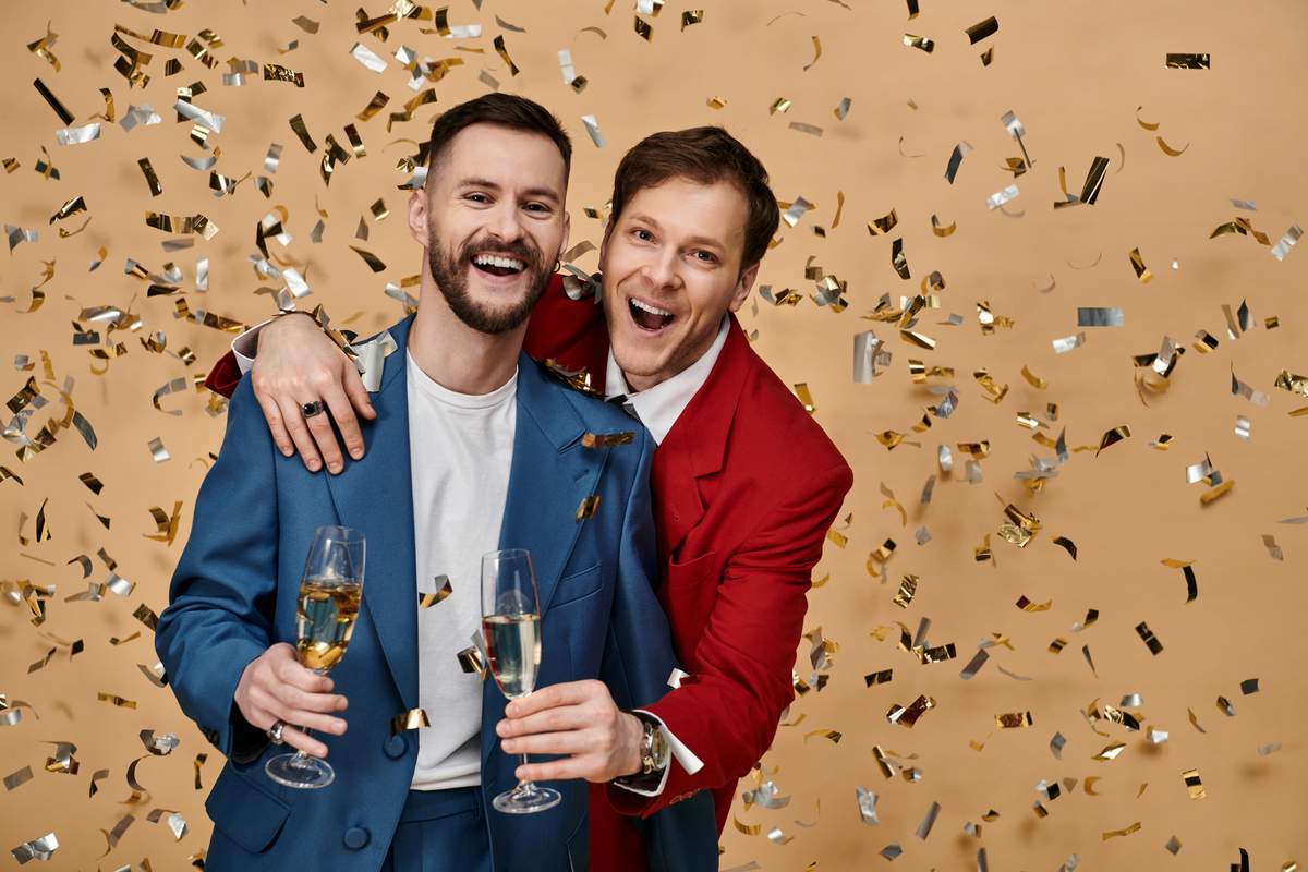 Two smiling men in suits, one with his arm around the other, holding champagne glasses as gold confetti falls