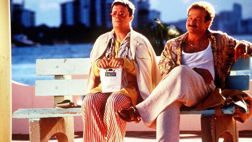 Nathan Lane and Robin Williams sitting on a bench by the water in colorful 1990s outfits in The Birdcage