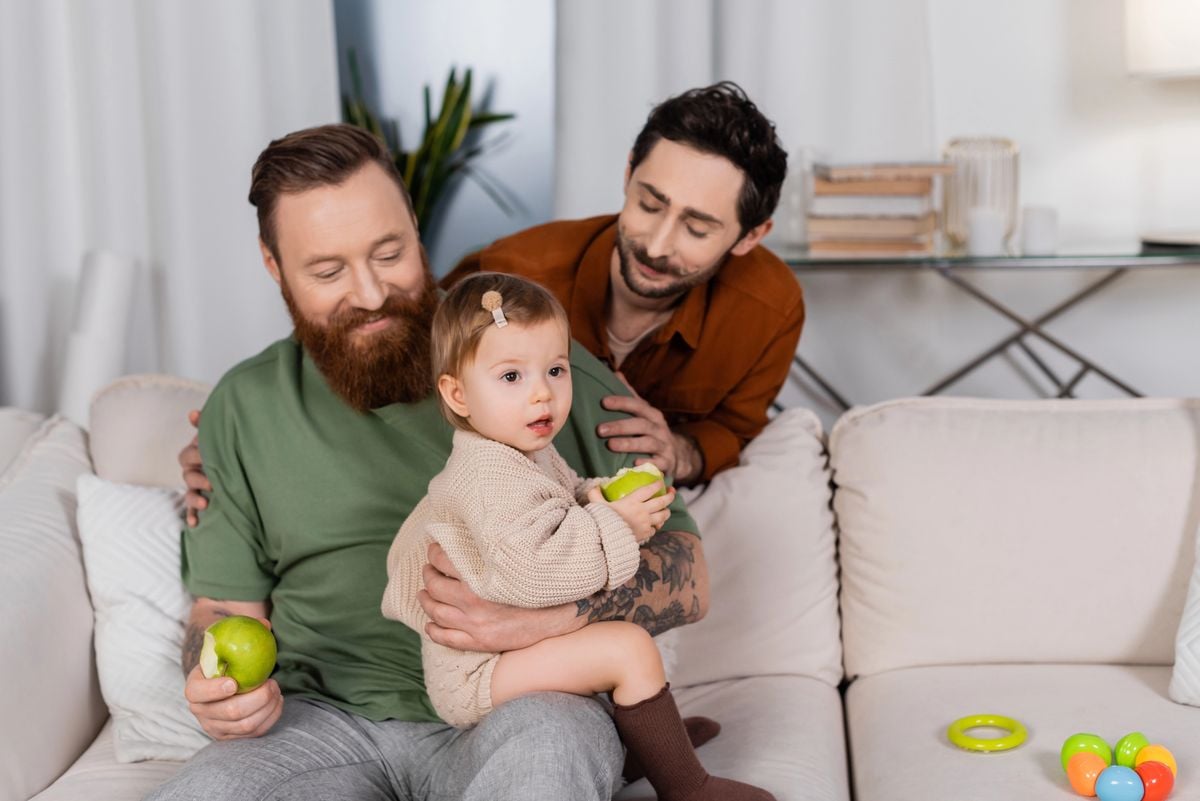 Two dads on a sofa smiling at their baby daughter, who sits on one dad's lap holding an apple
